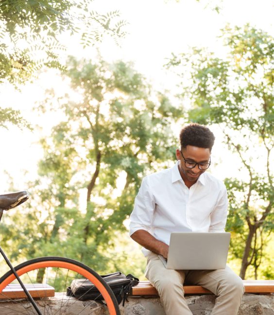 Picture of happy young african handsome man early morning with bicycle outdoors. Looking aside using laptop computer.